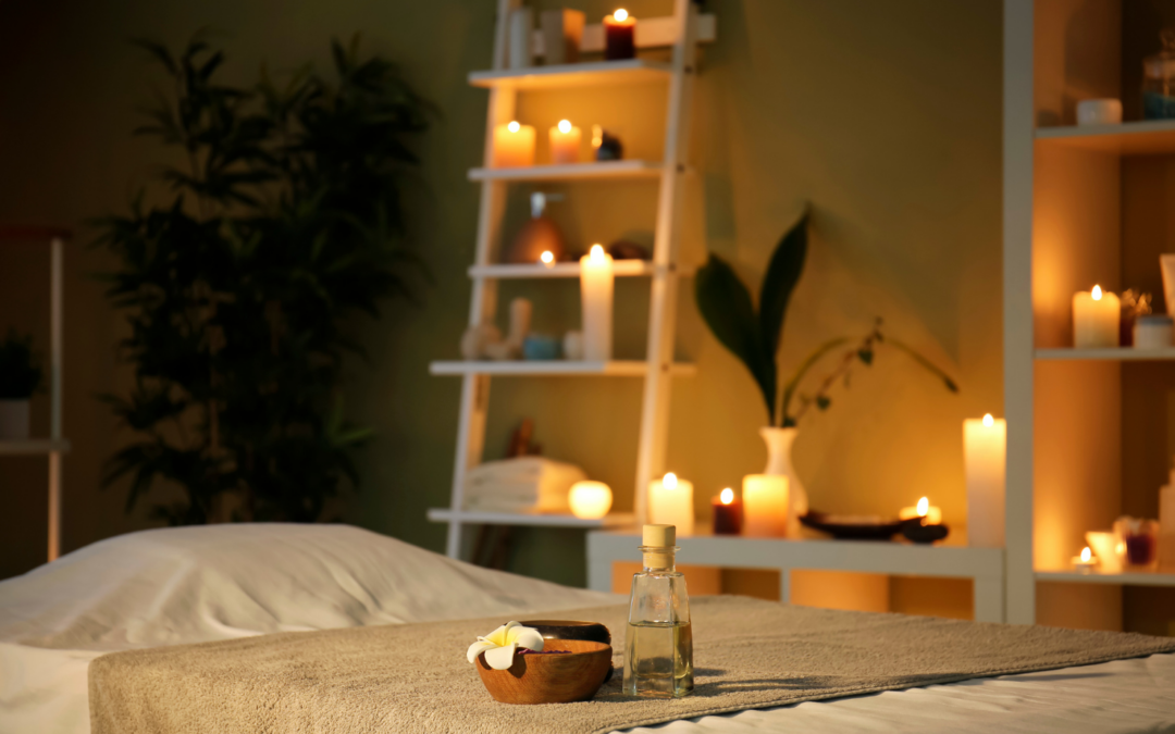 A softly lit massage room with a massage table covered in neutral linens. A small wooden bowl with a flower and a glass bottle of oil sit on the table. Shelves in the background are filled with glowing candles, plants, and spa decor, creating a warm and relaxing atmosphere for a holiday stress relief massage